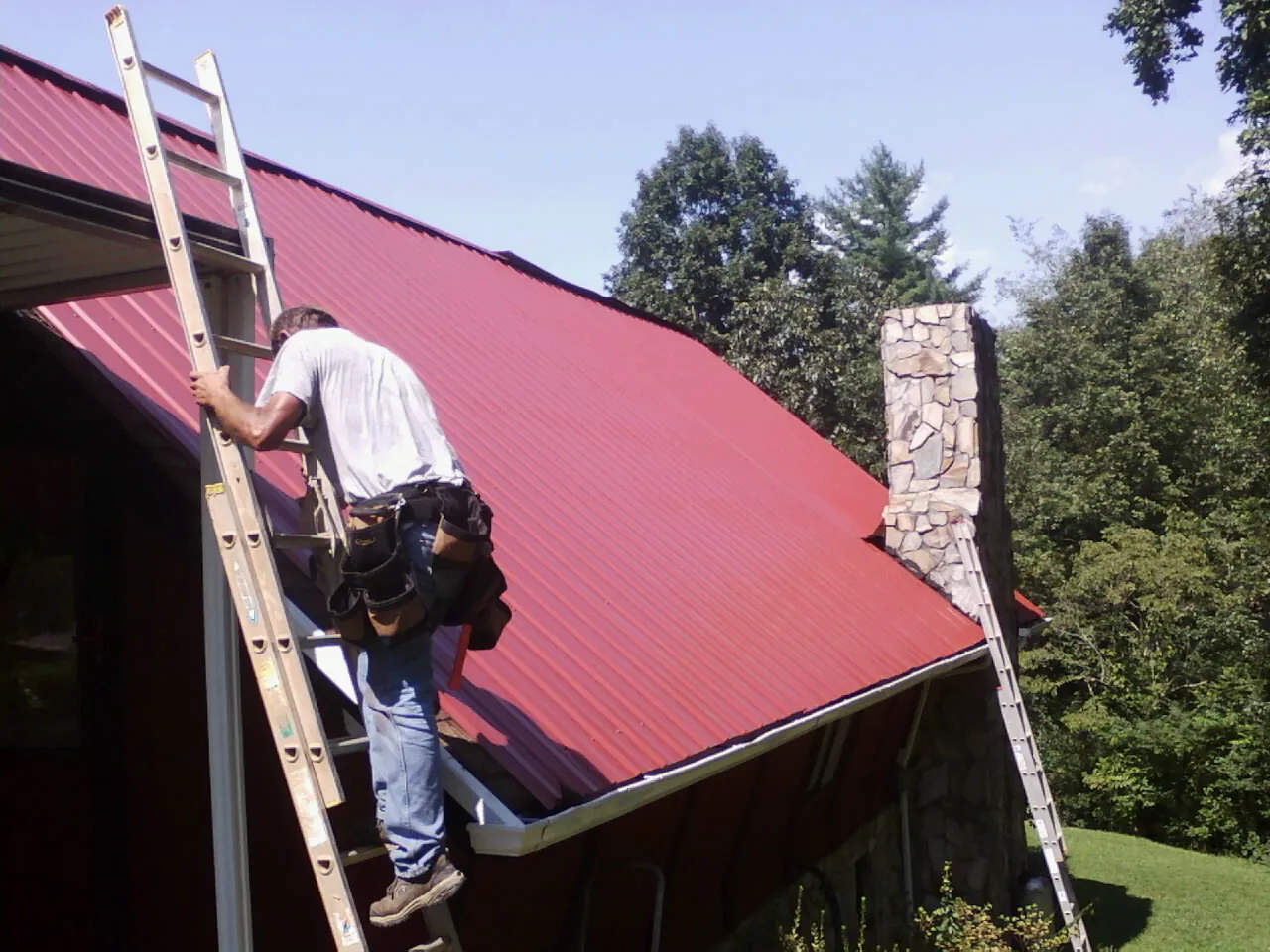 Roofer on a ladder beside a red metal roof and stone chimney — Soffit & Fascia Repair by Roofing Brothers in Westcliffe